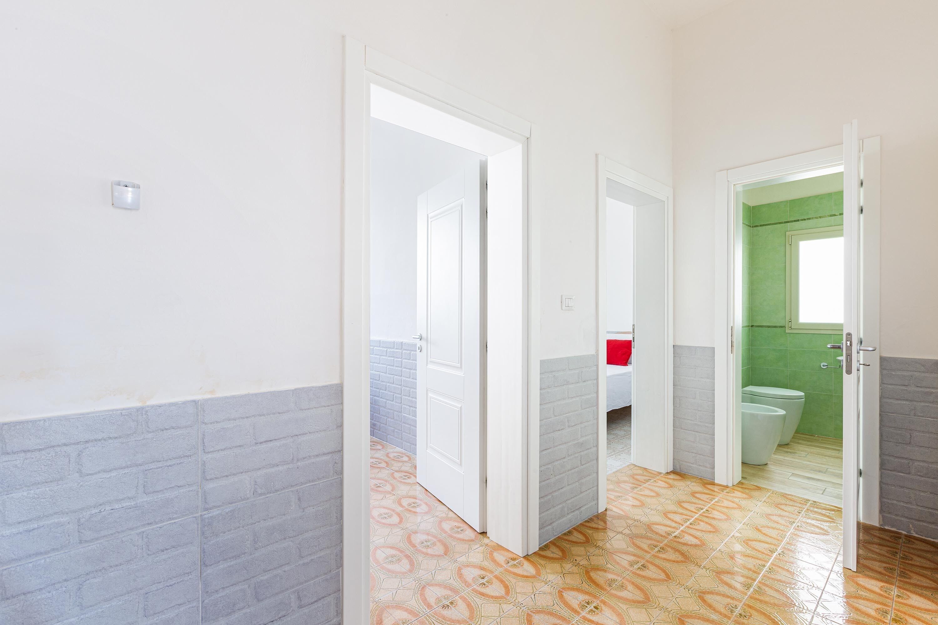 A hallway with a patterned floor, white walls, and a green-tiled bathroom visible through an open door.