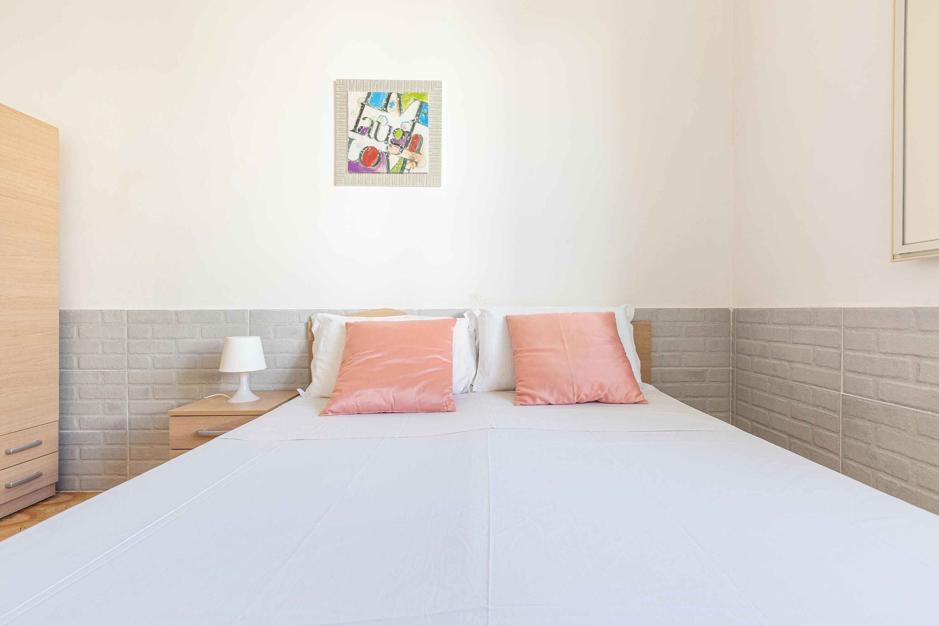 Una cama cuidadosamente tendida con almohadas blancas y rosas, una mesita de noche con una lámpara, y un cuadro abstracto de colores en la pared.