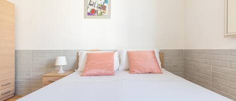 Una cama cuidadosamente tendida con almohadas blancas y rosas, una mesita de noche con una lámpara, y un cuadro abstracto de colores en la pared.