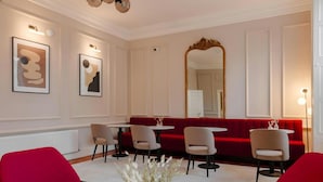 Un comedor con una banca tapizada en rojo, paredes blancas y un espejo grande. Hay cuatro sillas alrededor de una mesa pequeña, y un jarrón con flores en el suelo.