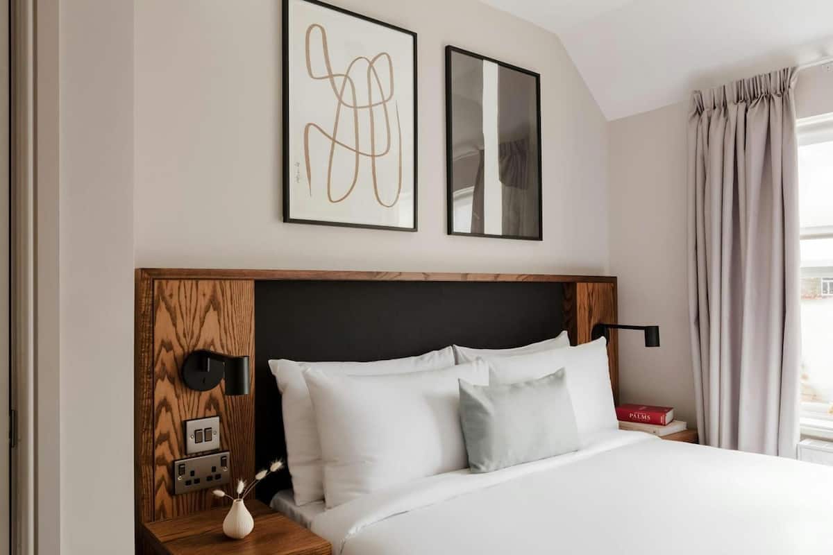 Un dormitorio moderno con cabecera de madera, ropa de cama blanca, una mesita de noche con lámpara y dos cuadros enmarcados en la pared.