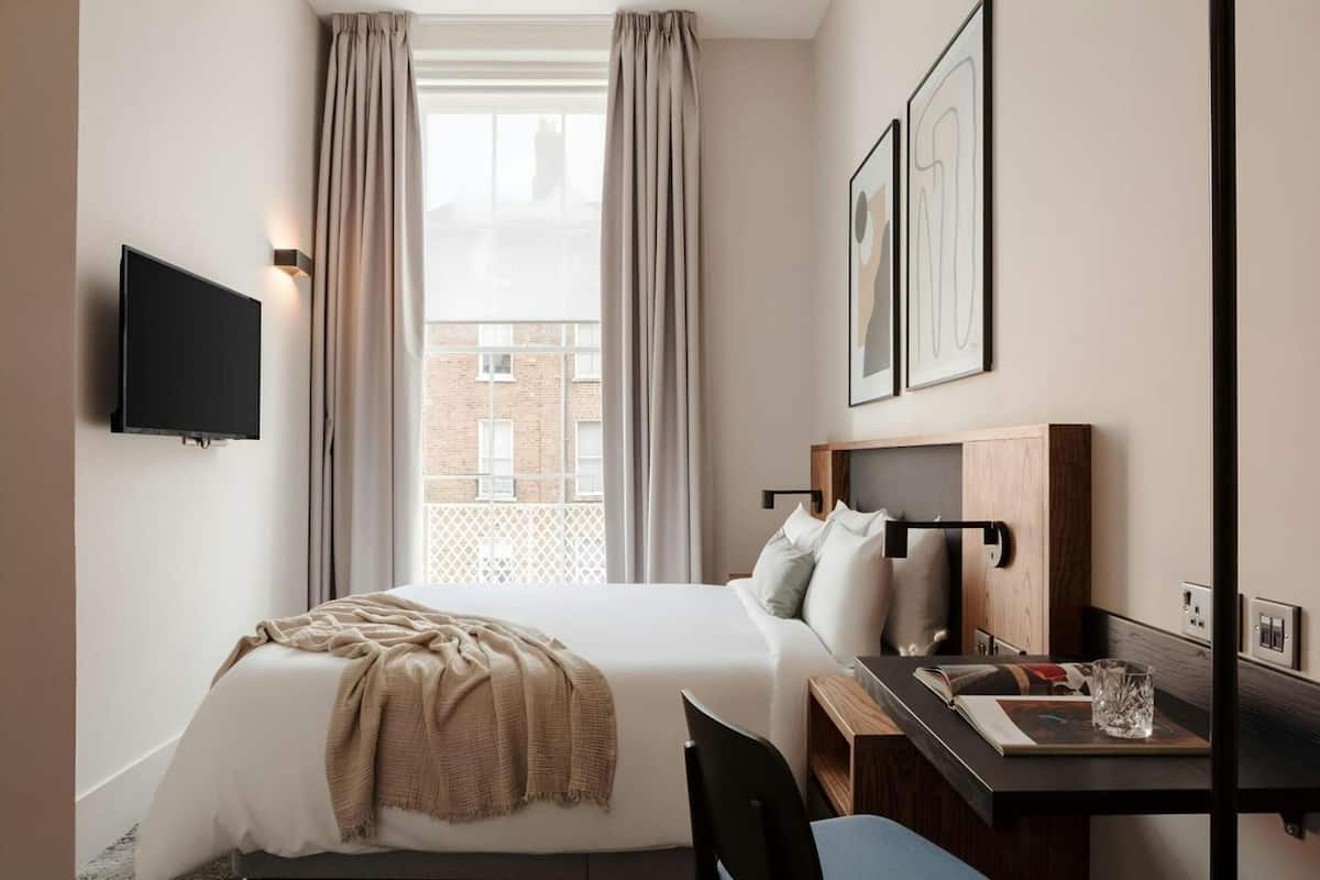 Habitación de hotel con cama, escritorio, silla, televisor montado en la pared y ventana con cortinas.