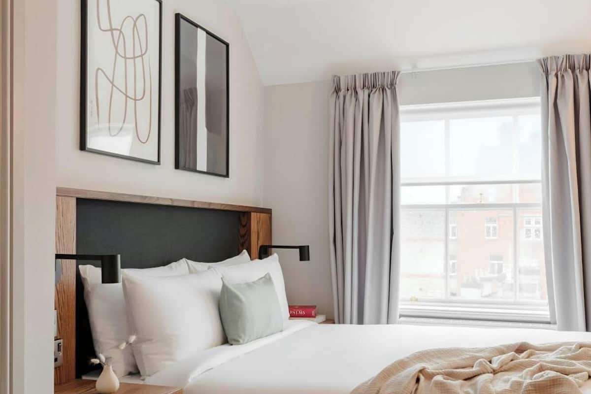 Una habitación de hotel con una cama grande, una mesita de noche, cortinas en la ventana y dos cuadros enmarcados en la pared.