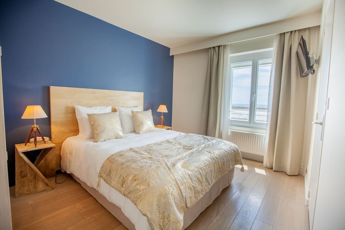Een slaapkamer met een groot bed, twee nachtkastjes met lampen, een raam met gordijnen en uitzicht op zee.