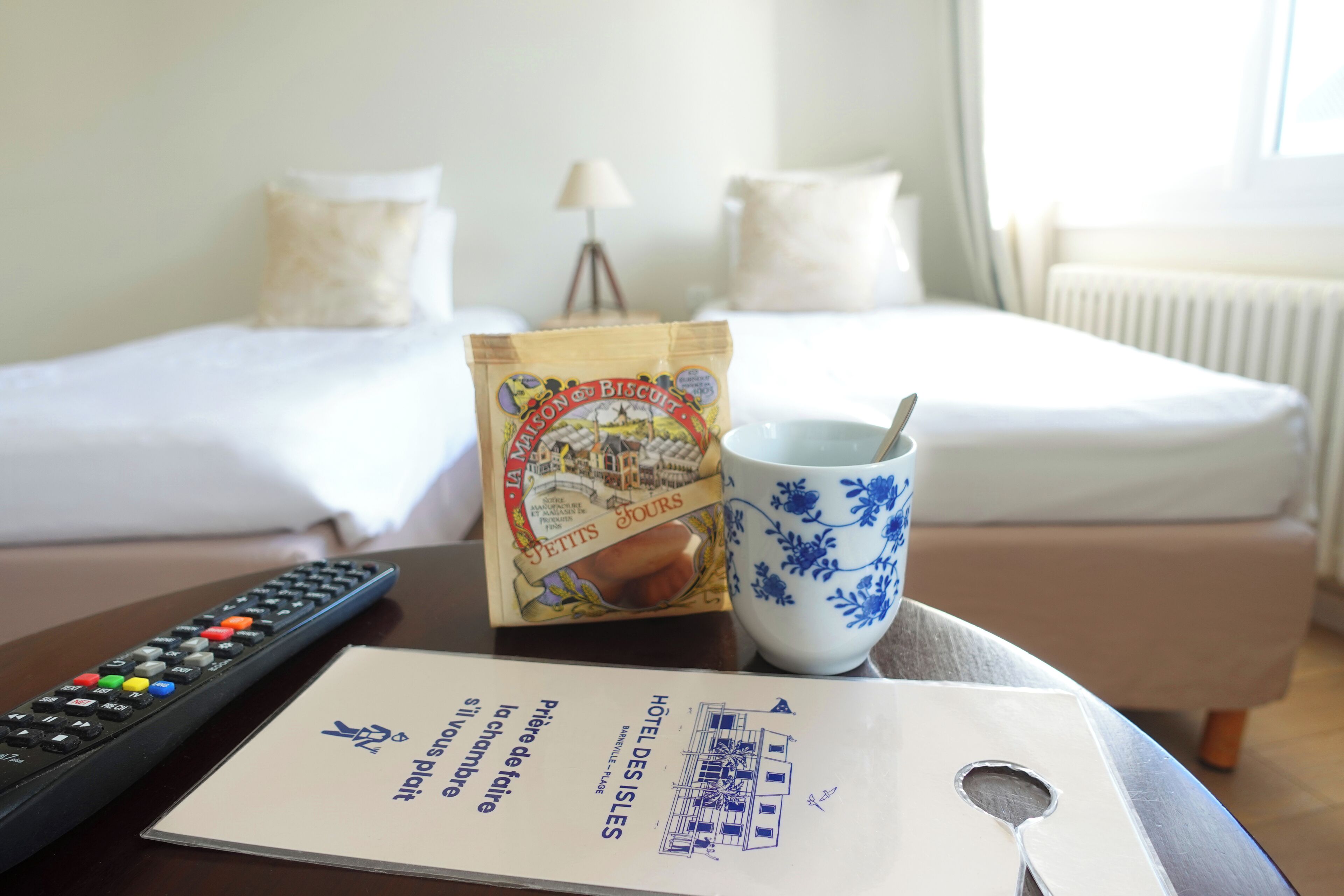 A hotel room with a bed, a remote control, a packet of biscuits, a blue and white mug, and a menu.