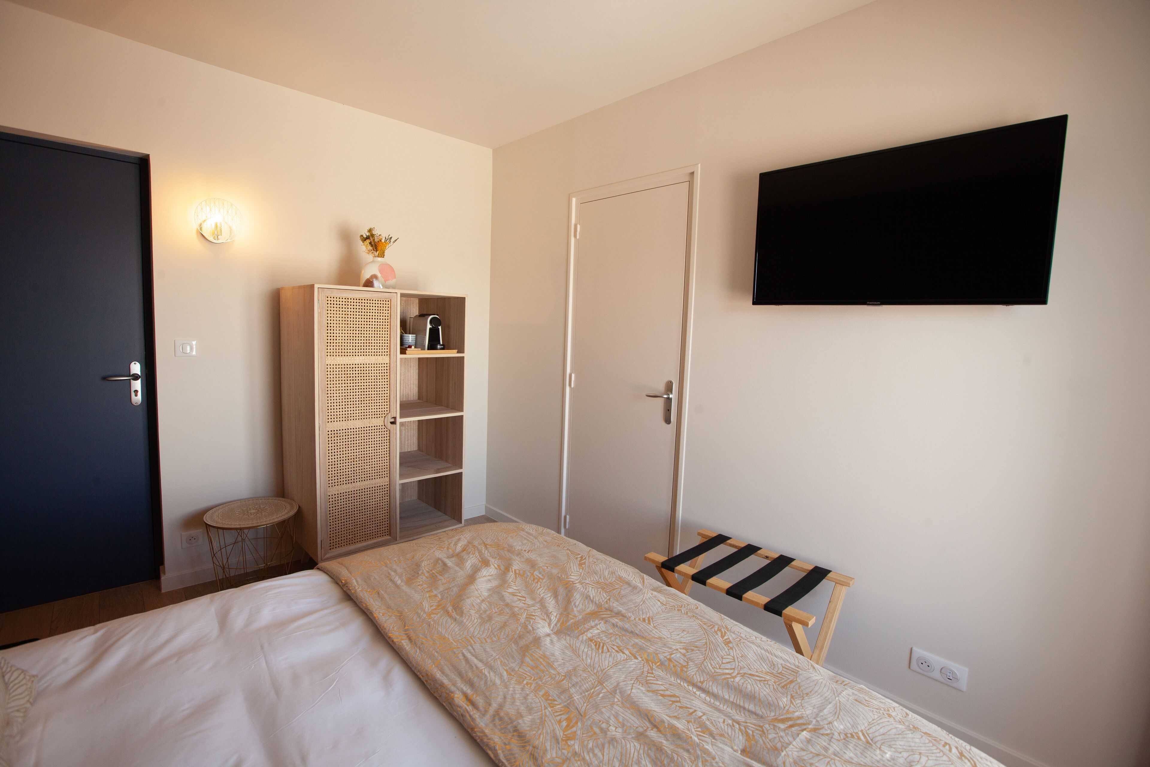 A bedroom with a bed, a wardrobe, a small table, a bench, a TV mounted on the wall, and a door.