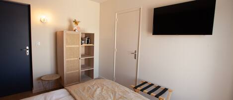 A bedroom with a bed, a wardrobe, a small table, a bench, a TV mounted on the wall, and a door.