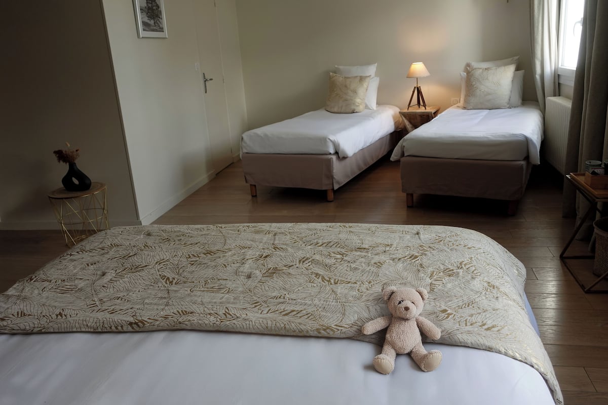Een hotelkamer met twee enkele bedden, een groot bed met decoratieve plaid, een teddybeer op het grote bed, een nachtkastje met een lamp en een kleine tafel met een vaas.