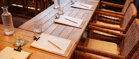 A wooden dining table set with glassware, napkins, and cutlery, surrounded by wicker chairs with cushioned seats.