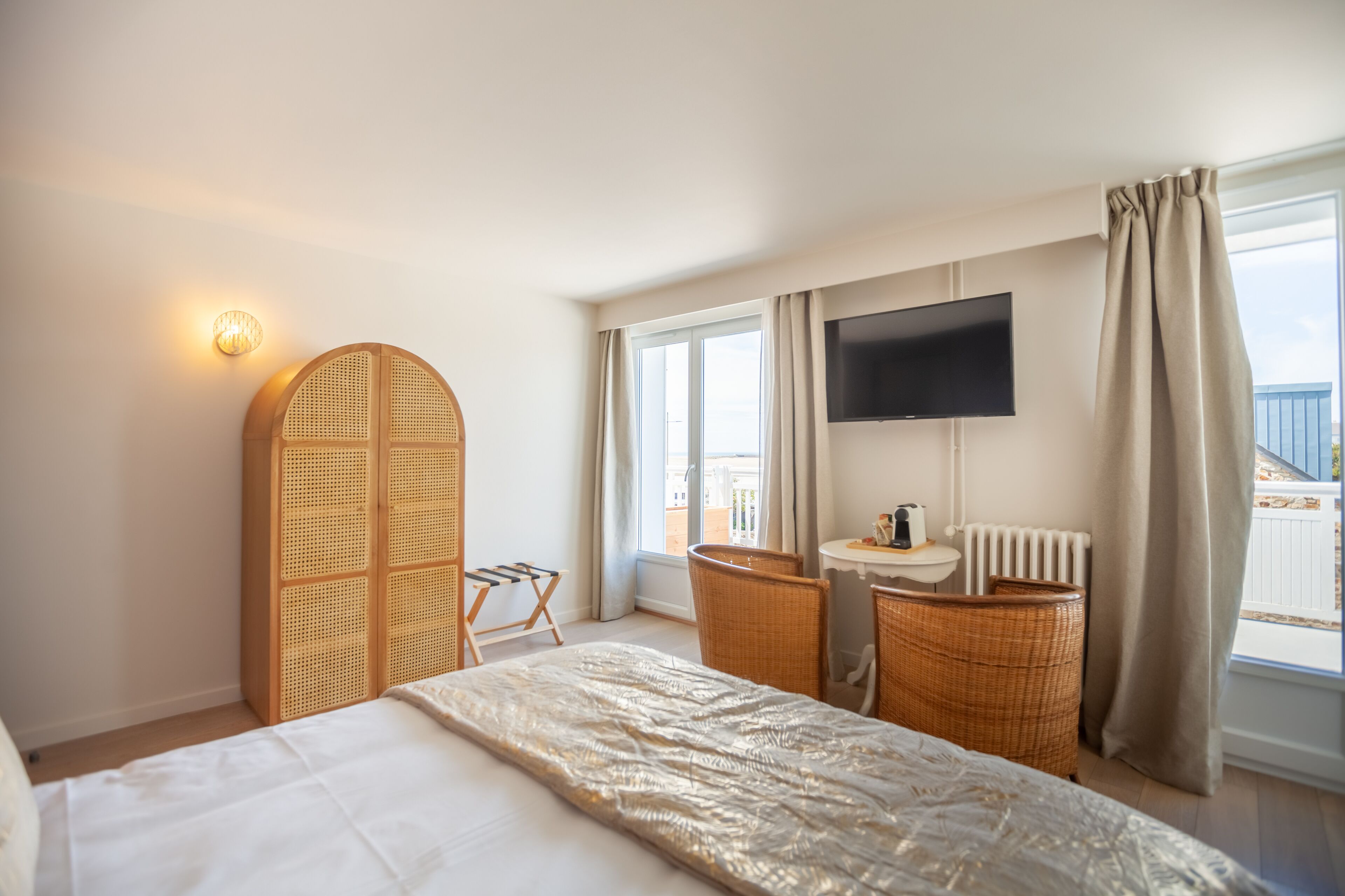 A hotel room with a bed, a television, a small table, a chair, a wicker chair, and a view of the outside.