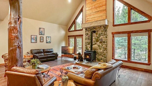 Ein geräumiges Wohnzimmer mit Kamin aus Stein, Ledersofas und großen Fenstern mit Blick auf den Wald.