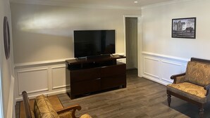 A living room with a flat-screen TV on a wooden stand, a patterned armchair, and a framed picture on the wall.