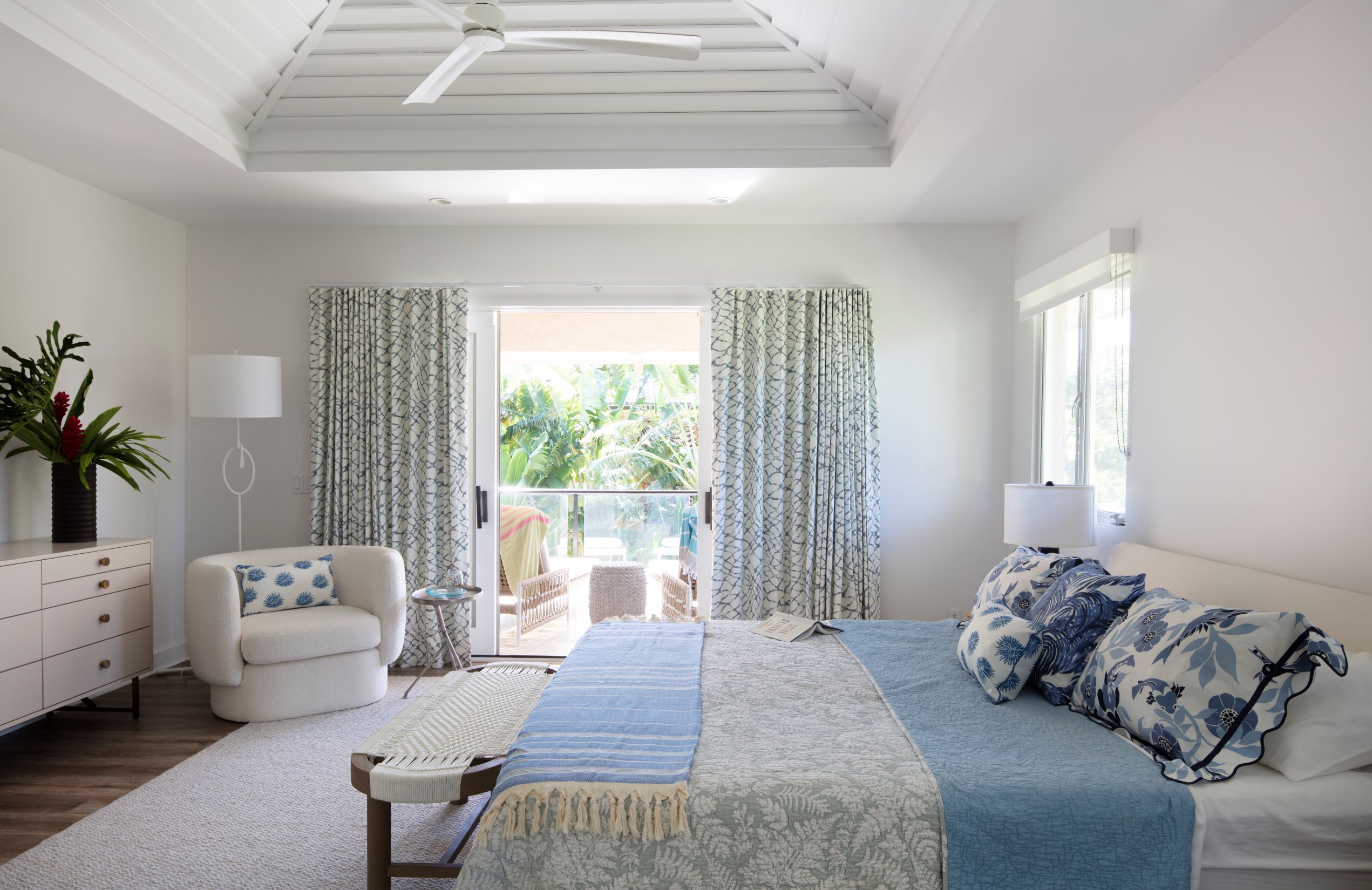 A bedroom with a bed, a chair, a small table, a lamp, and a view of the outdoors.