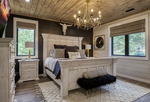 A bedroom with a large bed, a chandelier, a cow skull decoration, and a window with a view of trees.