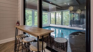 A pool area with a wooden table and stools, large windows, and a view of trees. - Summit Trails Mansion - Luxury Cabin with Views! (Sevierville)