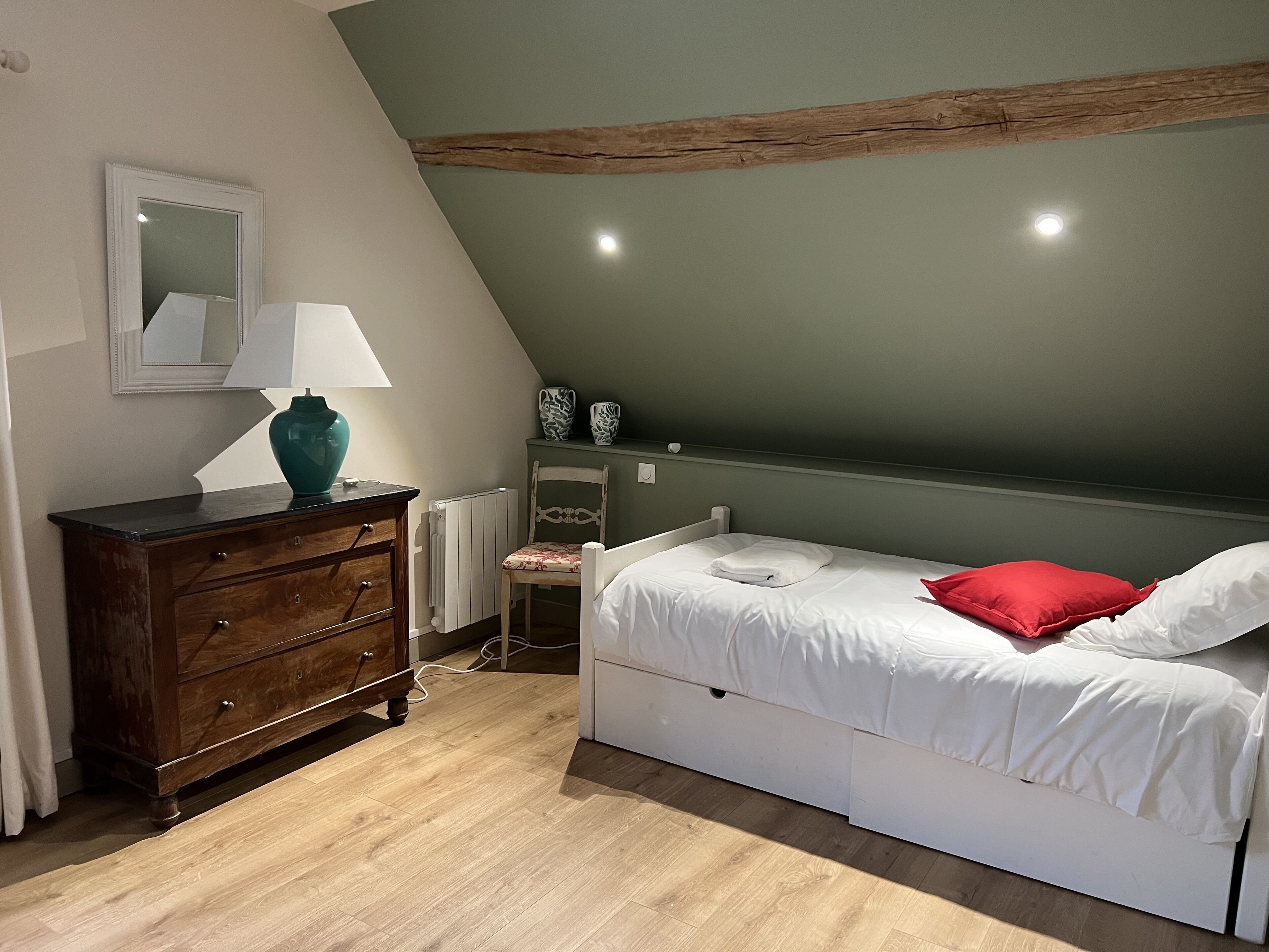 A bedroom with a sloped ceiling, a wooden dresser, a bed with white bedding and a red pillow, a mirror, and two decorative vases.