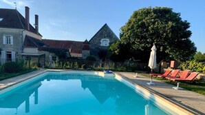 Una piscina rectangular con superficie de agua azul cristalina, rodeada por una terraza pavimentada. Hay sillas de descanso rojas y un paraguas blanco a la derecha. Se observa un árbol grande y un edificio de piedra con techo a dos aguas al fondo.