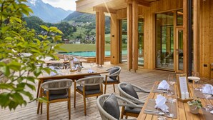 Una terraza de madera con mesas y sillas, con vistas a un paisaje montañoso y una piscina.