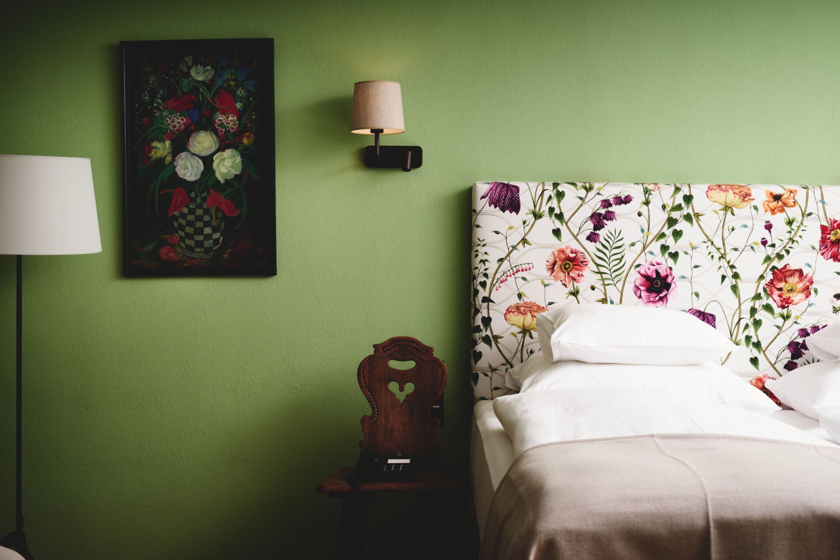 A neatly made bed with floral bedding, a wall-mounted lamp, and a painting of flowers on a green wall.