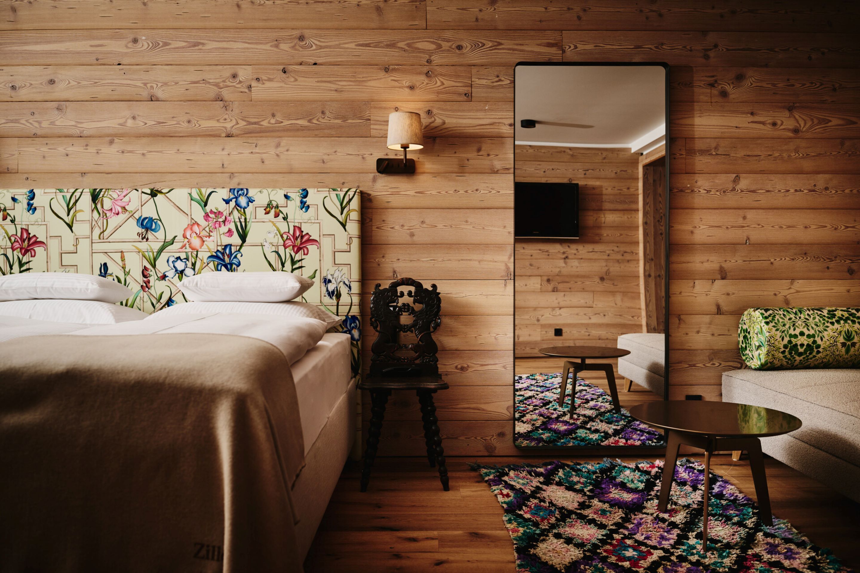 Habitación de hotel con pared de madera, cama con cabecera floral, una mesita, un sofá y un espejo.