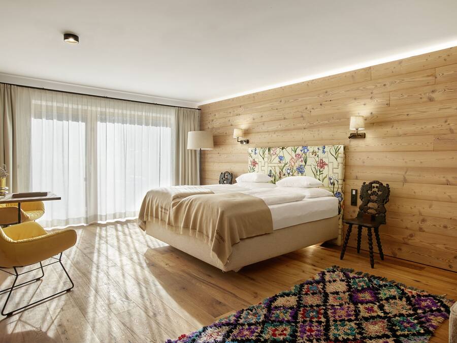A modern hotel room with a wooden floor, a large bed, a yellow armchair, a small table, and a colorful rug.
