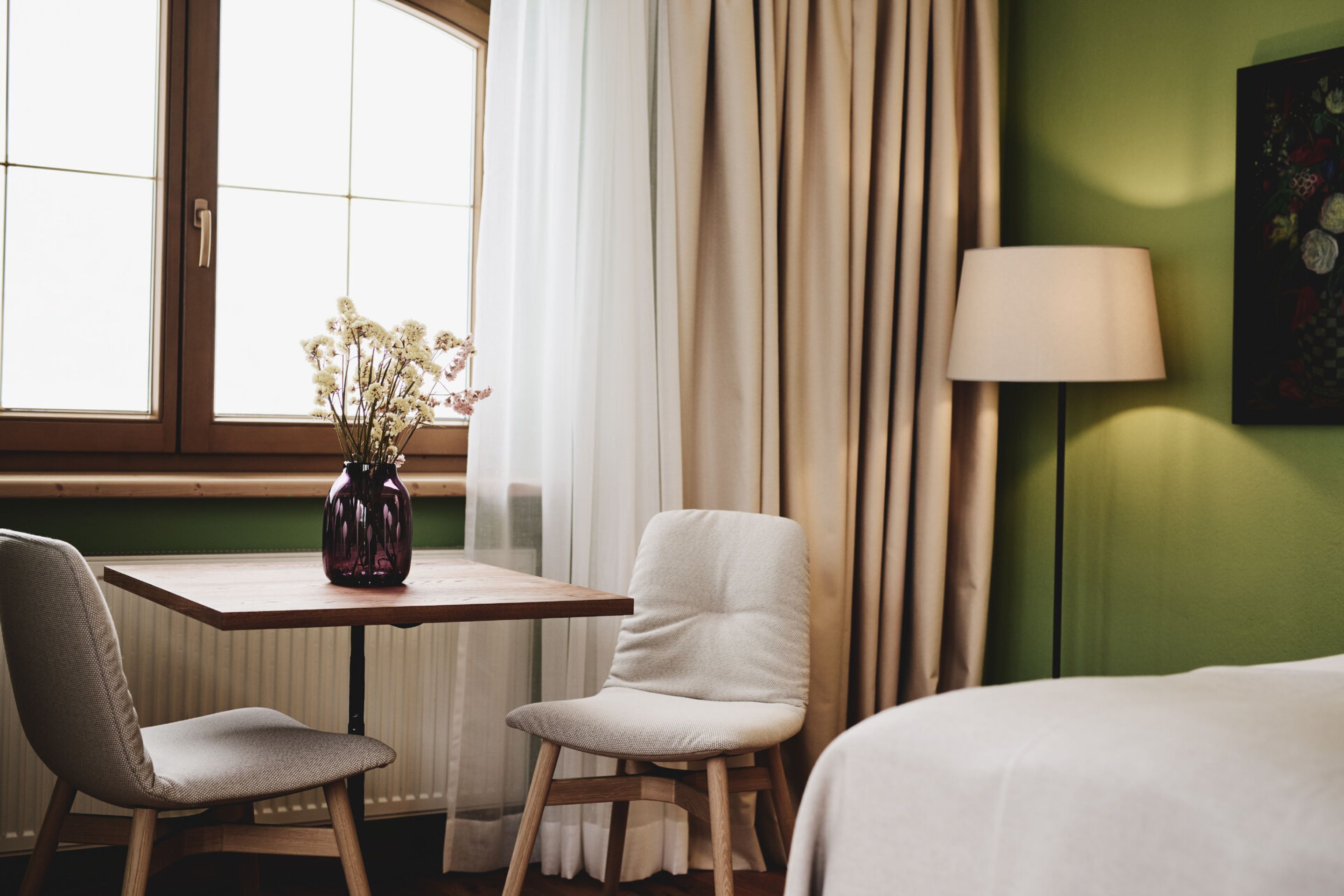 Una habitación de hotel con una mesa de madera, dos sillas, una cama, una lámpara y un ventanal con cortinas.