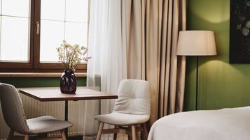 Una habitación de hotel con una mesa de madera, dos sillas, una cama, una lámpara y un ventanal con cortinas.
