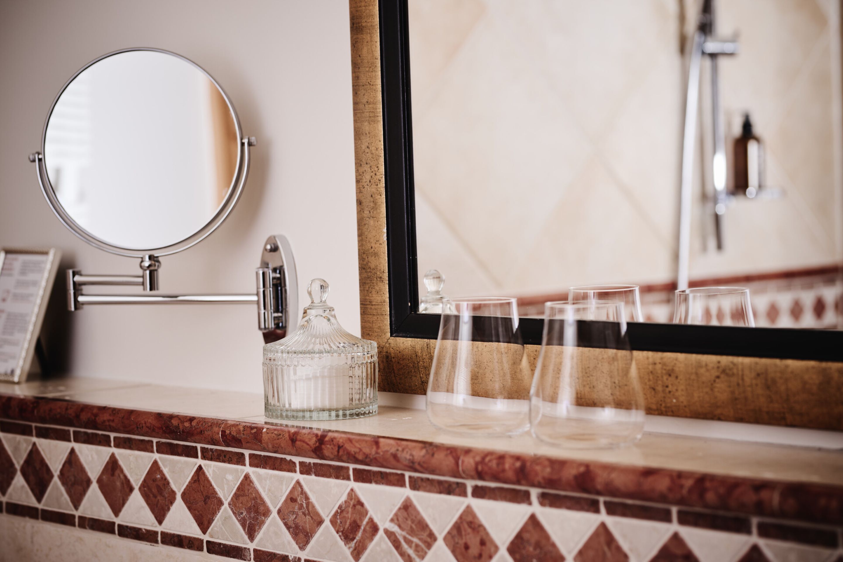 Un tocador de baño con un espejo redondo, una jarra de vidrio y tres vasos sobre la encimera.