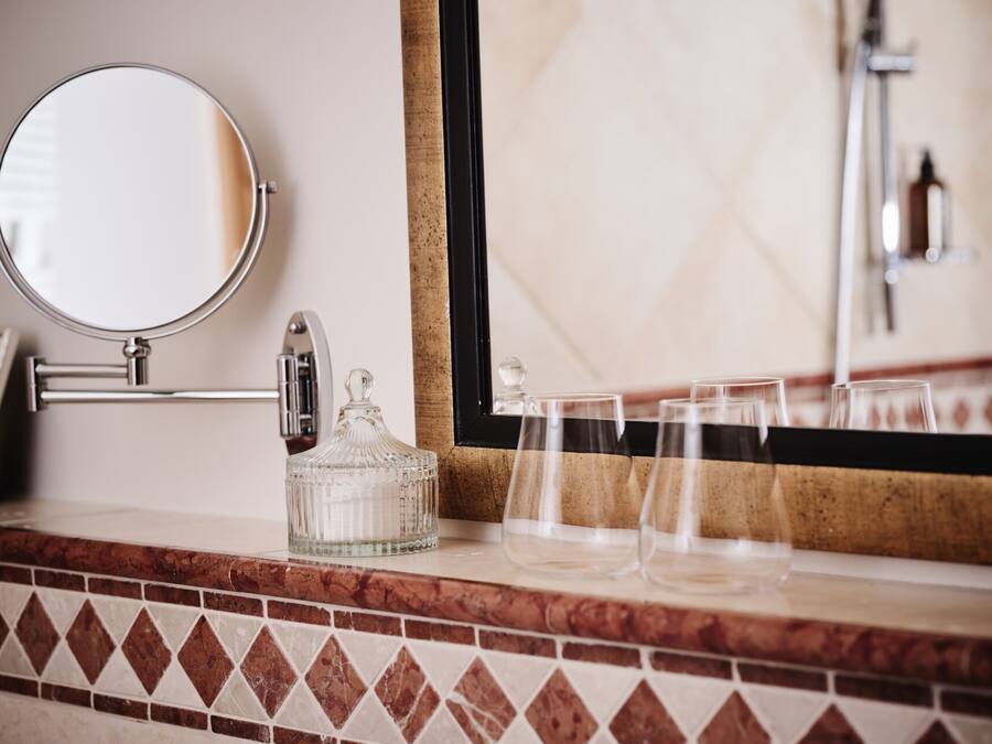 Un tocador de baño con un espejo redondo, una jarra de vidrio y tres vasos sobre la encimera.