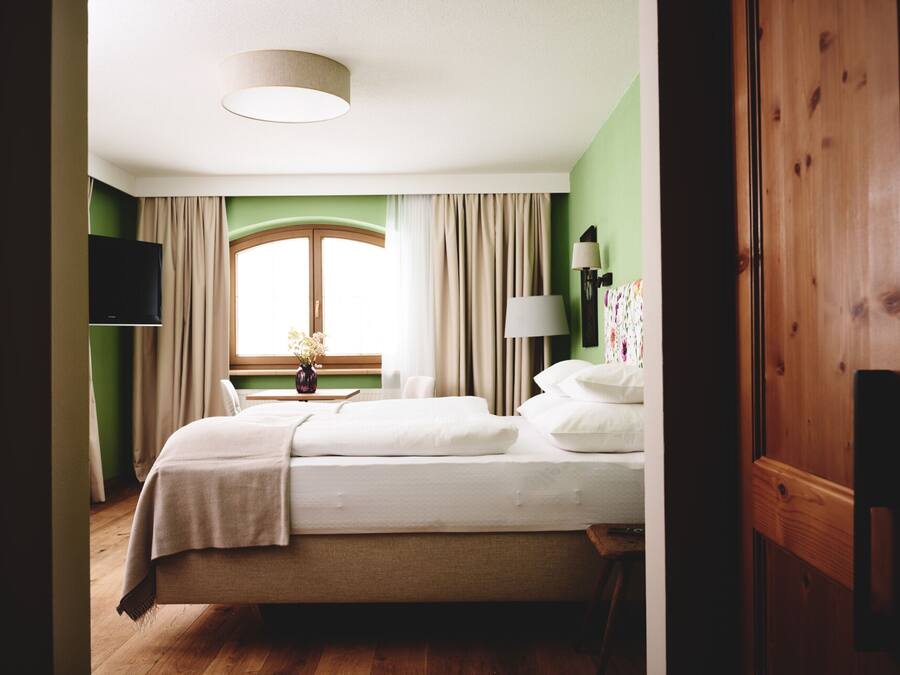 Un dormitorio ordenado con cama, ventana con cortinas, lámpara de noche y televisor de pantalla plana montado en la pared.