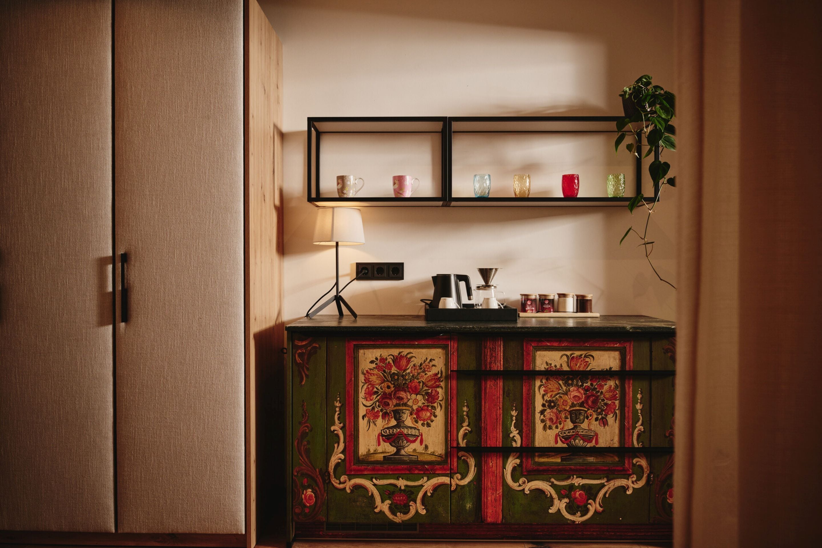 A green cabinet with floral designs, a shelf with cups and a plant, and a lamp.