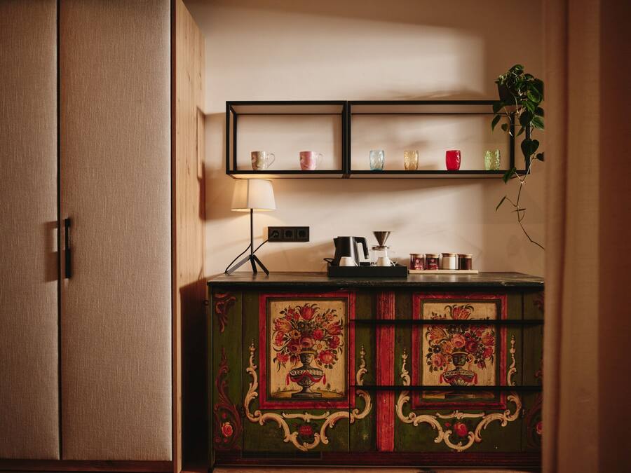 A green cabinet with floral designs, a shelf with cups and a plant, and a lamp.