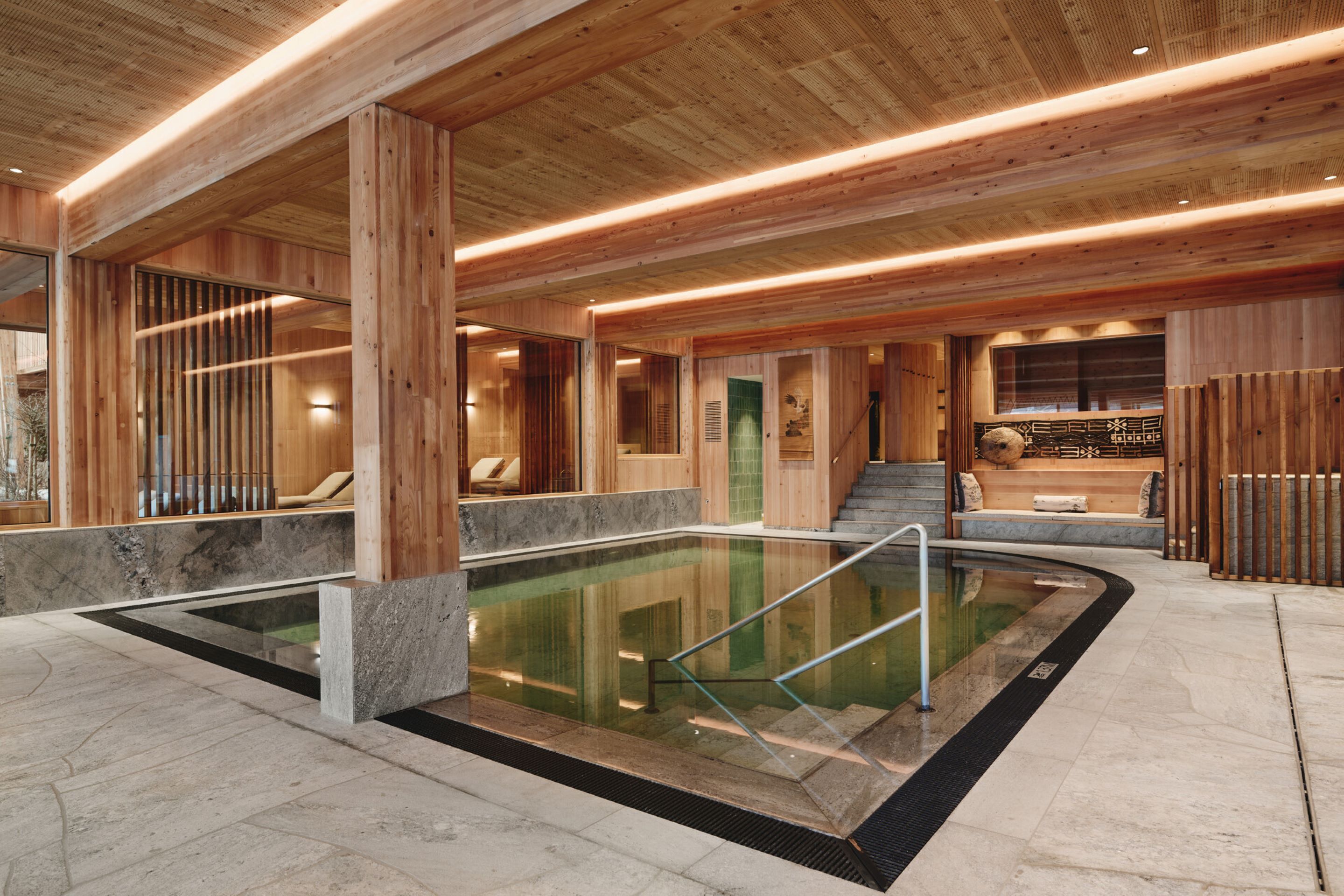 Un área de piscina interior moderna con vigas de madera, barandilla de vidrio y piso de piedra.