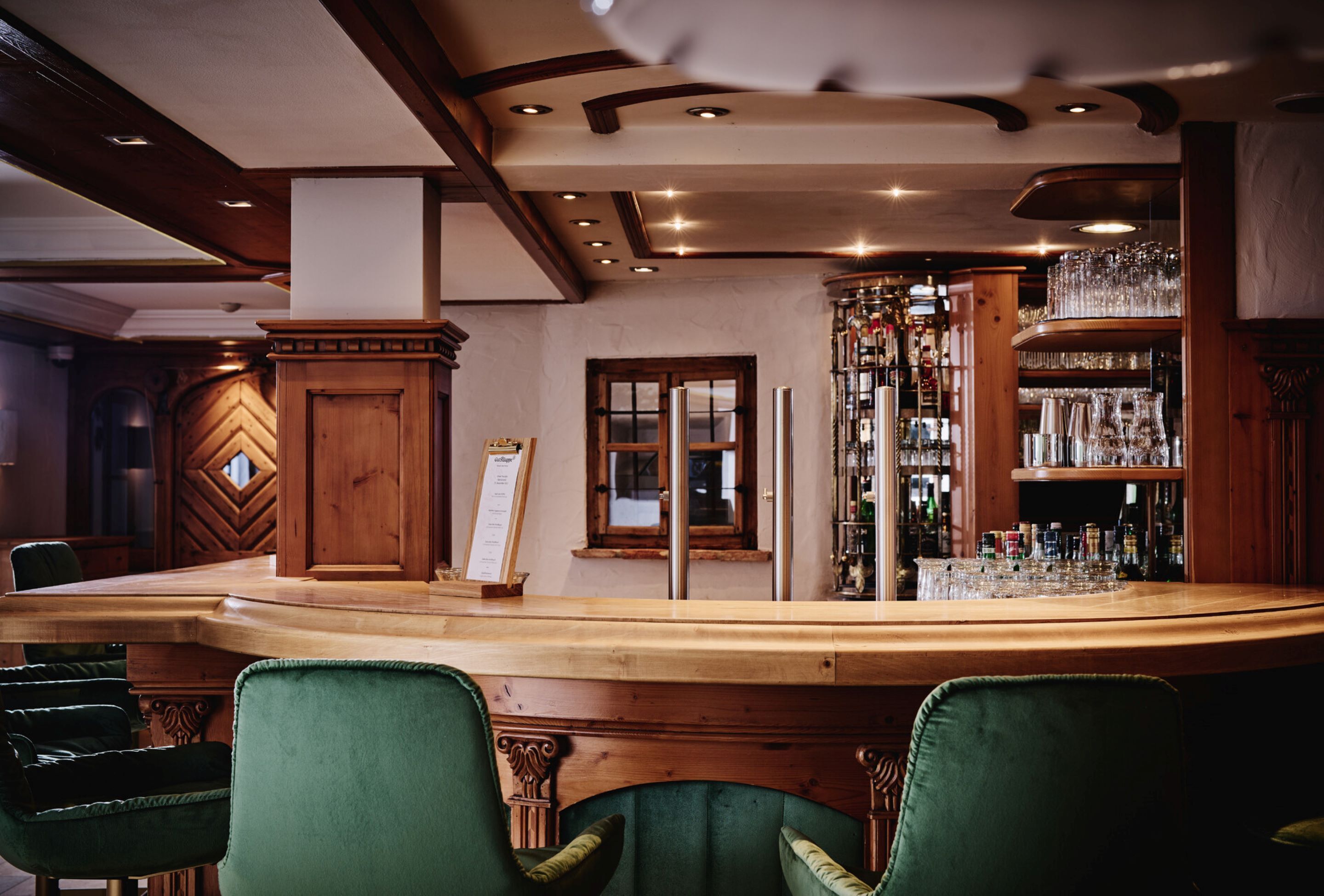 Een bar met een houten toonbank, groene bekleden stoelen en een glazen vitrine vol flessen.