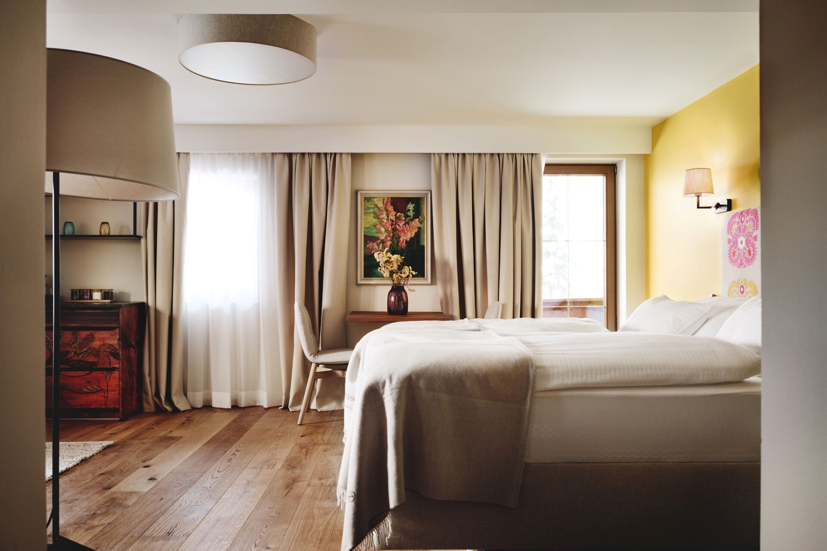 A hotel room with a bed, a desk with a chair, a lamp, a painting, and a window with curtains.