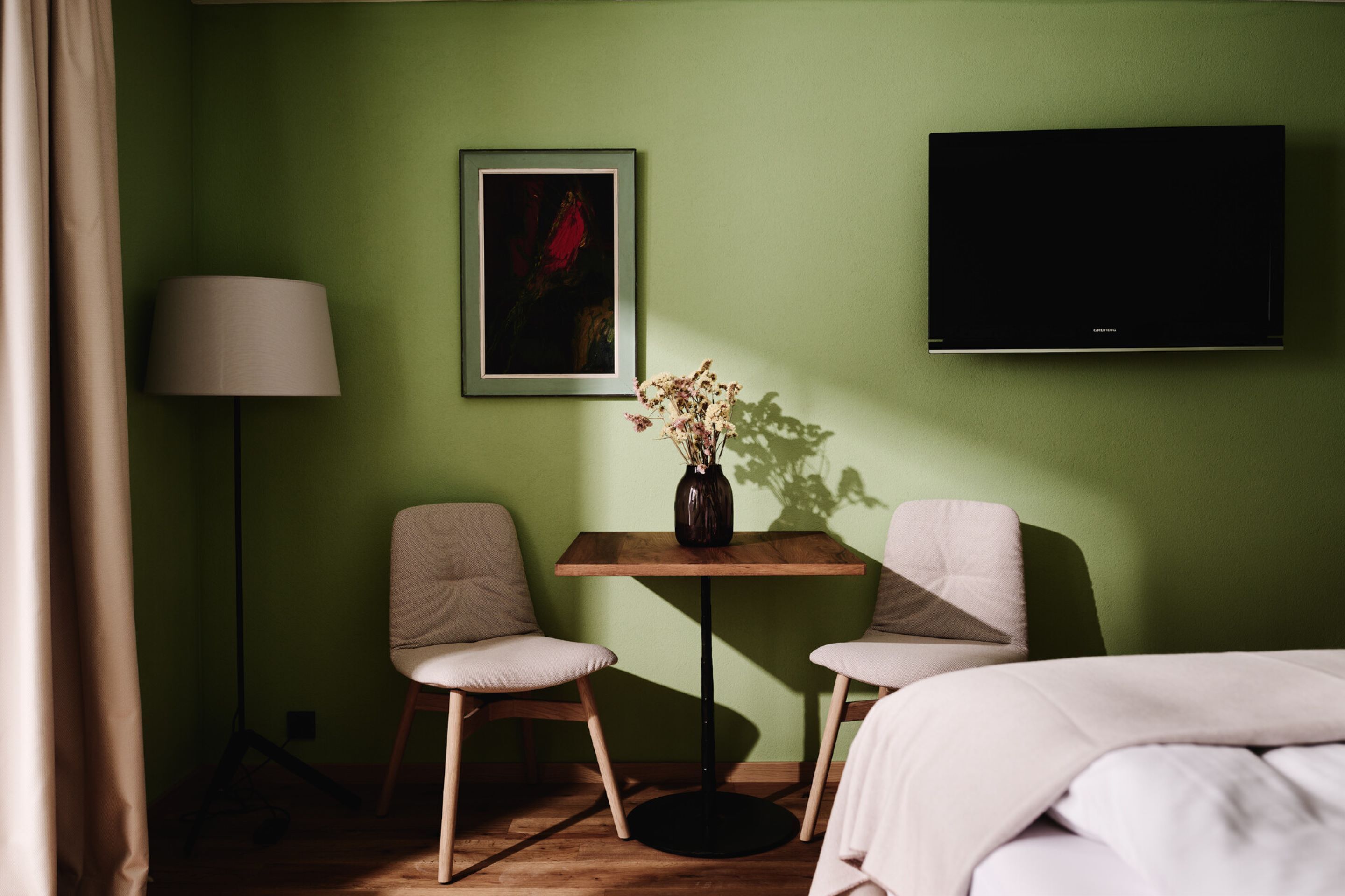 A hotel room with a bed, two chairs, a small table with a vase, a lamp, and a TV mounted on the wall.