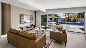 A modern living room with a large TV, comfortable seating, and a view of a swimming pool. - Modern Posh – Mid-Century Oasis with Pool, Spa, Game Yard & Mountain Views (Palm Springs)