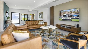 A modern living room with a brown leather sofa, a coffee table, and a TV mounted on the wall. - Modern Posh – Mid-Century Oasis with Pool, Spa, Game Yard & Mountain Views (Palm Springs)