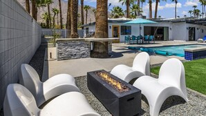 A pool area with white lounge chairs, a fire pit, and a poolside building with outdoor seating. - Modern Posh – Mid-Century Oasis with Pool, Spa, Game Yard & Mountain Views (Palm Springs)