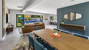 A modern living room with a wooden dining table, blue chairs, a sofa, and a large window overlooking a pool. - Modern Posh – Mid-Century Oasis with Pool, Spa, Game Yard & Mountain Views (Palm Springs)