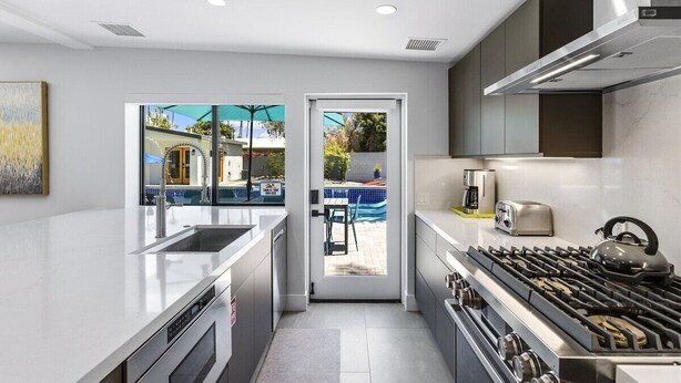 A modern kitchen with a large island, built-in oven, and gas stove. There is a view of a pool through the glass door. - Modern Posh – Mid-Century Oasis with Pool, Spa, Game Yard & Mountain Views (Palm Springs)