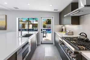 Une cuisine moderne dotée d’un grand îlot central, d’un four encastré et d’une cuisinière à gaz. On peut admirer une piscine à travers la porte vitrée.