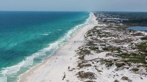 Ein Luftbild eines Strandes mit freiem Blick auf den Ozean, Sandküste und einer Mischung aus Vegetation und Gebäuden entlang der Küste.