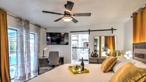 A hotel room with a large bed, a desk, a chair, a ceiling fan, a TV, and a view of the pool.