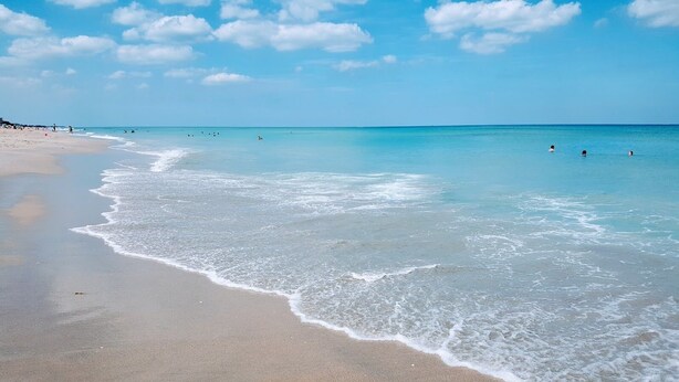 Une plage avec une eau d’un bleu limpide, de douces vagues et quelques personnes qui se baignent.