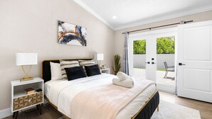 Une chambre à coucher comprenant un lit, des tables de chevet, un tableau et une vue sur l’extérieur, accessible par une porte coulissante qui donne sur un espace extérieur.