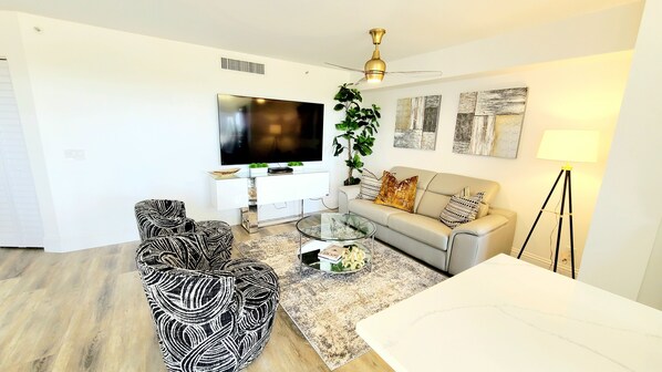 A modern living room with a flat-screen TV, a glass coffee table, a grey sofa, and a black and white patterned armchair. - POSHPADZ -OceanView Villa-Resort Complex, Penthouse, Sleeps 4, Walk to Beach (Jupiter)