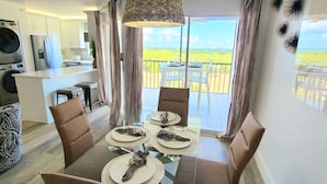 A dining area with a table set for four, a kitchen in the background, and a balcony with a view. - POSHPADZ -OceanView Villa-Resort Complex, Penthouse, Sleeps 4, Walk to Beach (Jupiter)