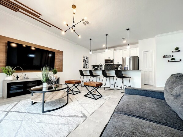A modern living room with a large TV, a dining area with bar stools, and a kitchen with white cabinets.