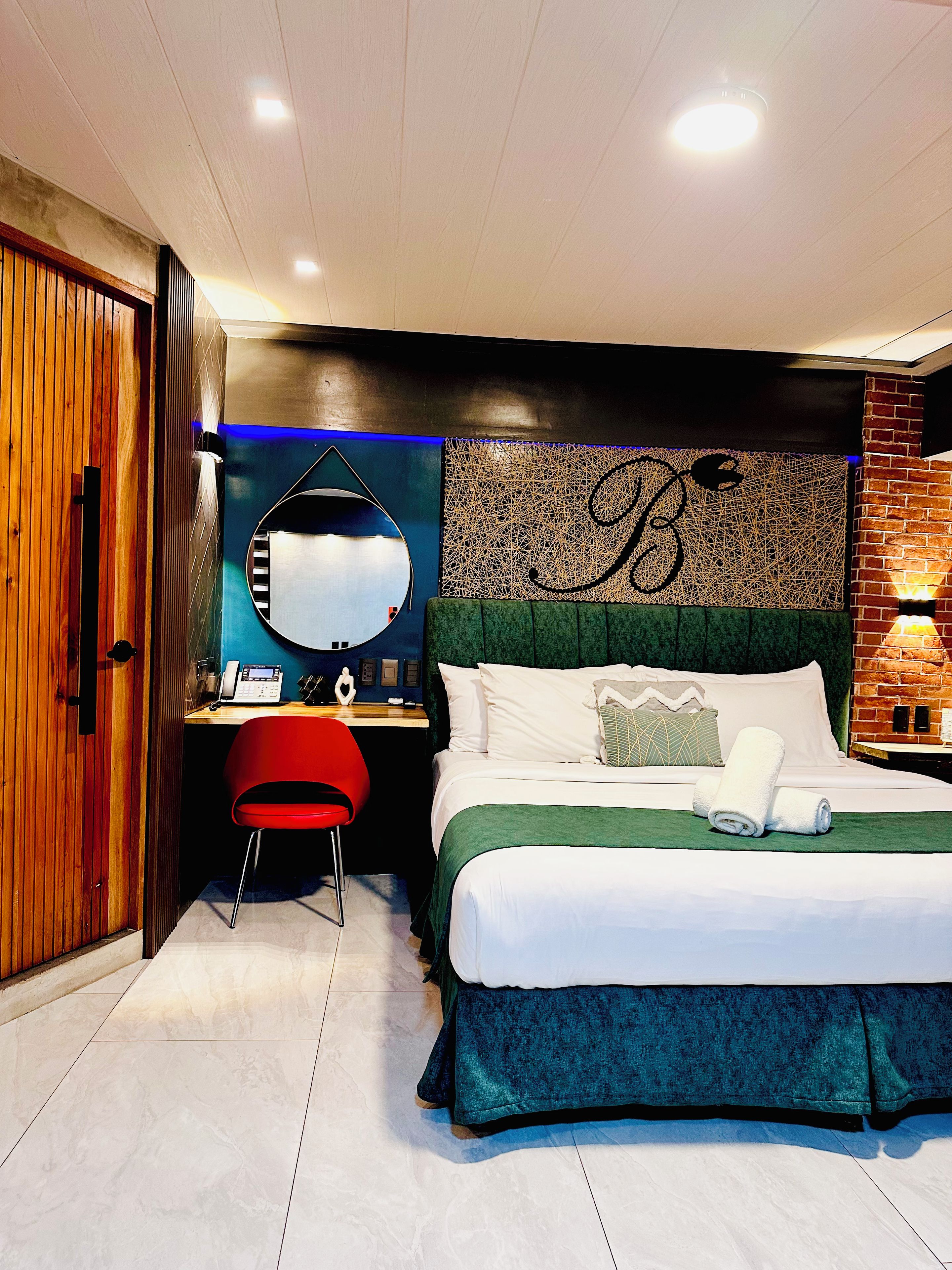 A modern hotel room with a large bed, a desk with a chair, a mirror, and a wall with a decorative pattern.